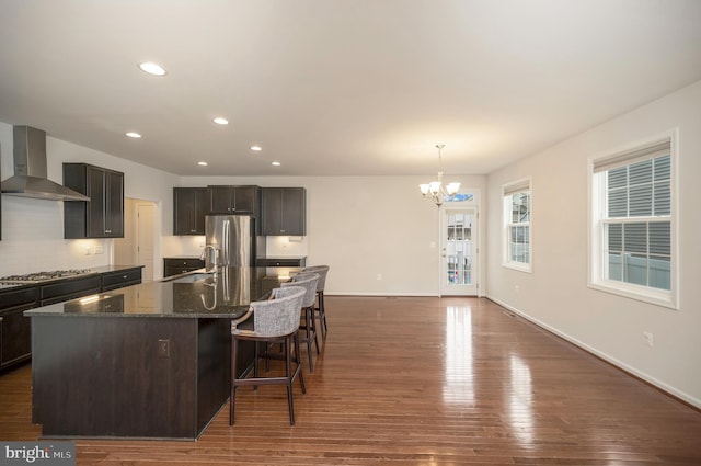 kitchen featuring wall chimney range hood, appliances with stainless steel finishes, a sink, dark stone countertops, and an inviting chandelier