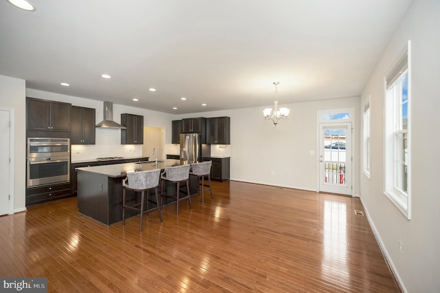 kitchen with wall chimney range hood, dark brown cabinets, a kitchen breakfast bar, a chandelier, and stainless steel appliances