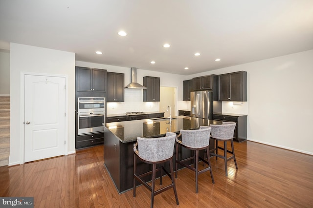kitchen featuring an island with sink, wall chimney exhaust hood, stainless steel appliances, a kitchen bar, and dark wood-type flooring