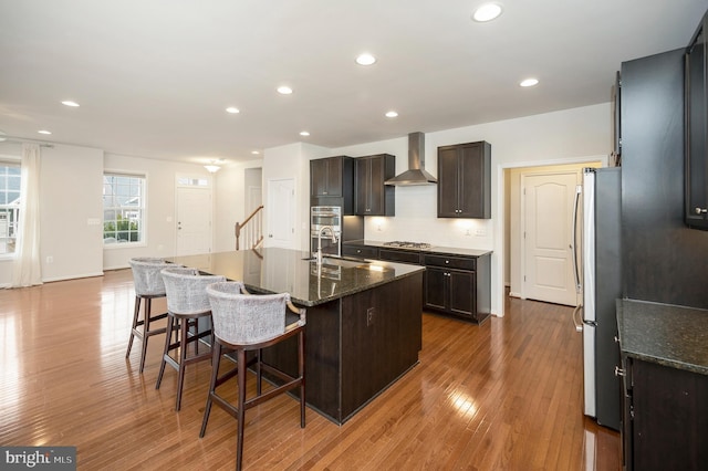 kitchen with wall chimney range hood, an island with sink, recessed lighting, wood finished floors, and a kitchen bar