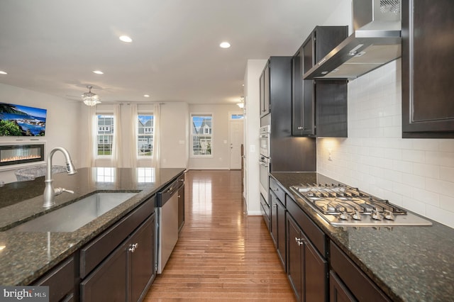kitchen featuring light wood-type flooring, wall chimney exhaust hood, a sink, backsplash, and stainless steel appliances