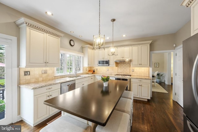 kitchen with an inviting chandelier, a sink, stainless steel appliances, cream cabinetry, and under cabinet range hood