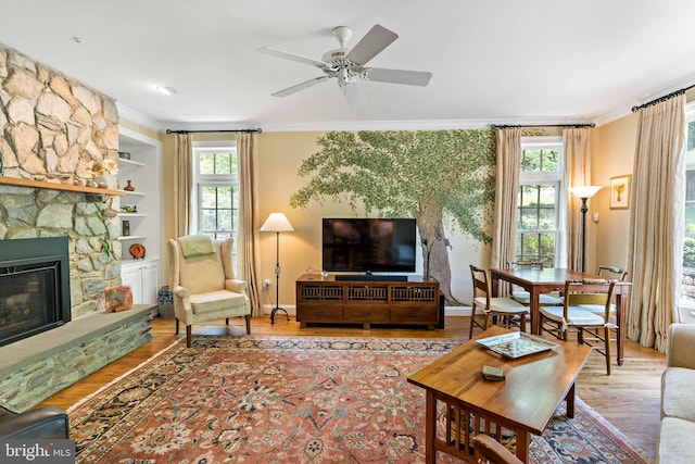 living area featuring crown molding, a ceiling fan, wood finished floors, and a stone fireplace