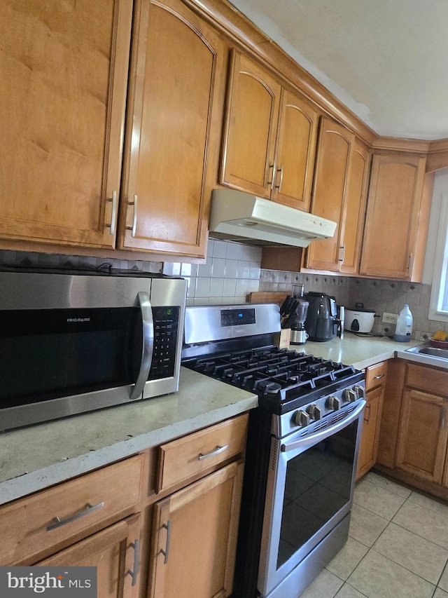 kitchen with under cabinet range hood, stainless steel appliances, brown cabinets, and backsplash