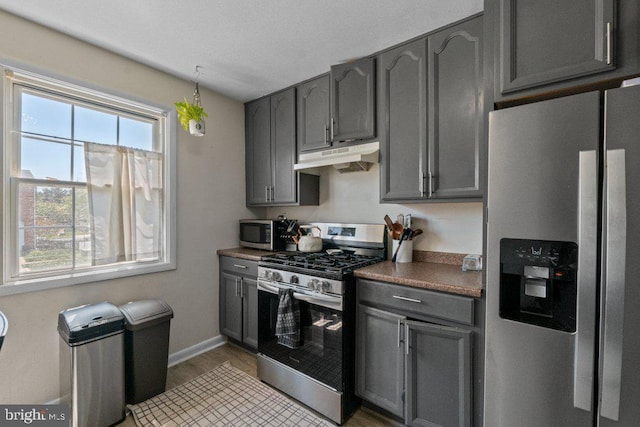 kitchen featuring appliances with stainless steel finishes, gray cabinetry, under cabinet range hood, dark countertops, and light wood finished floors