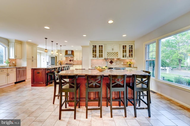 kitchen with cream cabinetry, wall chimney range hood, stainless steel dishwasher, a large island, and recessed lighting
