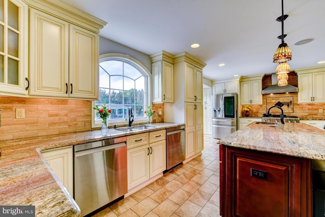 kitchen with cream cabinetry, stainless steel appliances, wall chimney exhaust hood, a sink, and light stone countertops