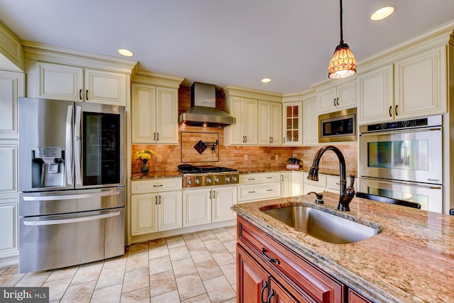 kitchen featuring appliances with stainless steel finishes, wall chimney exhaust hood, a sink, cream cabinets, and light stone countertops
