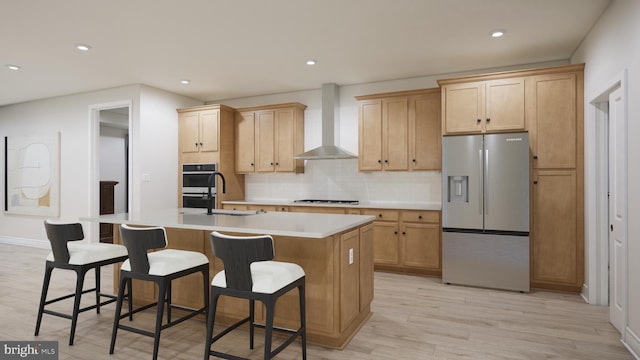 kitchen with appliances with stainless steel finishes, a sink, light wood-style floors, wall chimney exhaust hood, and an island with sink
