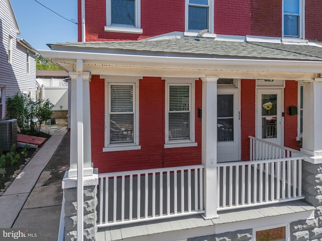 view of exterior entry with a porch, brick siding, cooling unit, and roof with shingles