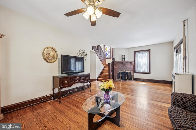 living area featuring wood-type flooring, a brick fireplace, stairs, a ceiling fan, and baseboards