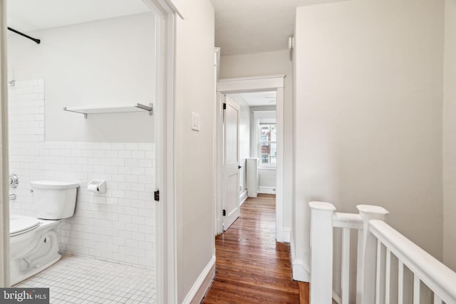 corridor with an upstairs landing, wood finished floors, wainscoting, and tile walls