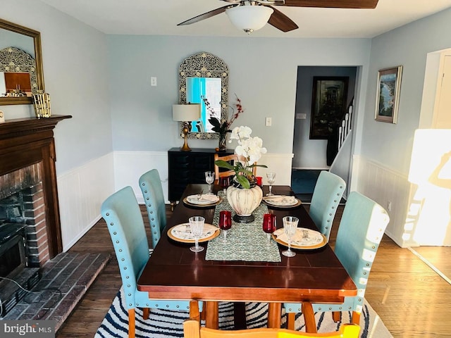 dining space featuring wood finished floors, a wainscoted wall, a brick fireplace, ceiling fan, and stairs