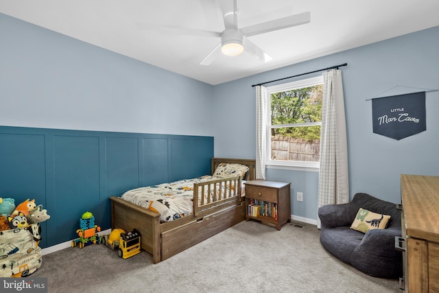 carpeted bedroom featuring visible vents, wainscoting, a decorative wall, ceiling fan, and a nursery area