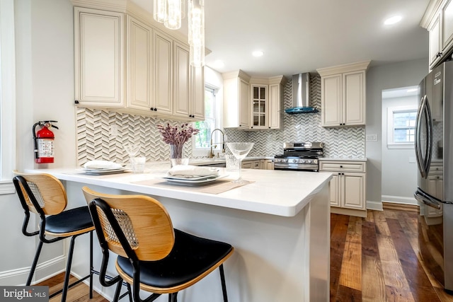 kitchen featuring wall chimney range hood, appliances with stainless steel finishes, a breakfast bar area, a peninsula, and cream cabinetry