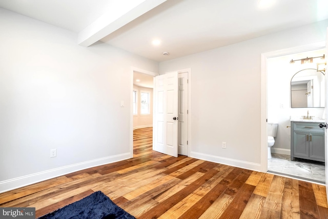 unfurnished bedroom with beamed ceiling, baseboards, connected bathroom, a sink, and light wood-style flooring