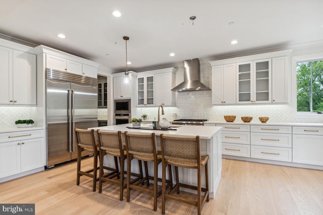 kitchen with built in appliances, an island with sink, wall chimney range hood, light countertops, and tasteful backsplash