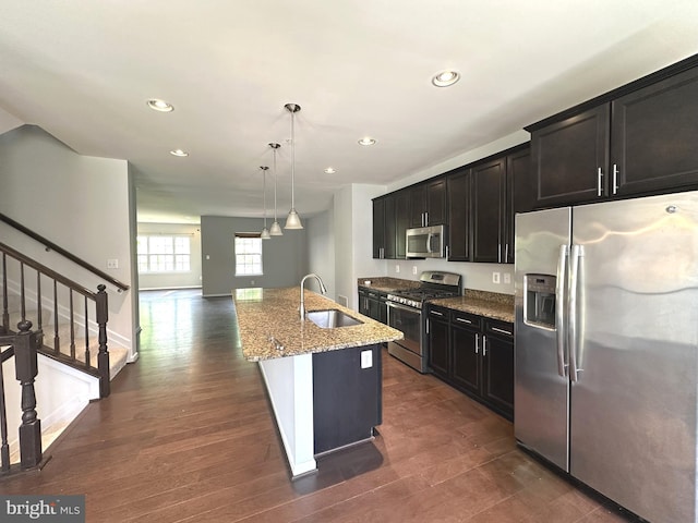kitchen featuring appliances with stainless steel finishes, a sink, light stone countertops, a center island with sink, and dark wood finished floors