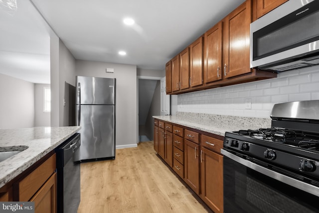 kitchen featuring brown cabinets, light wood finished floors, decorative backsplash, light stone countertops, and appliances with stainless steel finishes