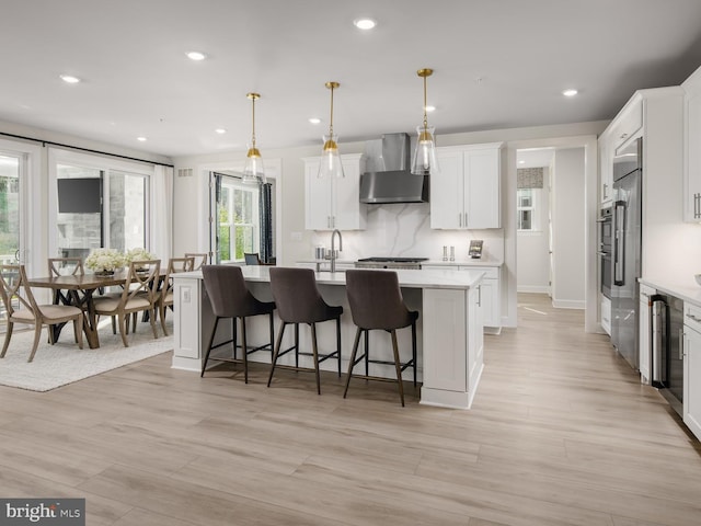 kitchen with light countertops, an island with sink, white cabinets, and wall chimney range hood