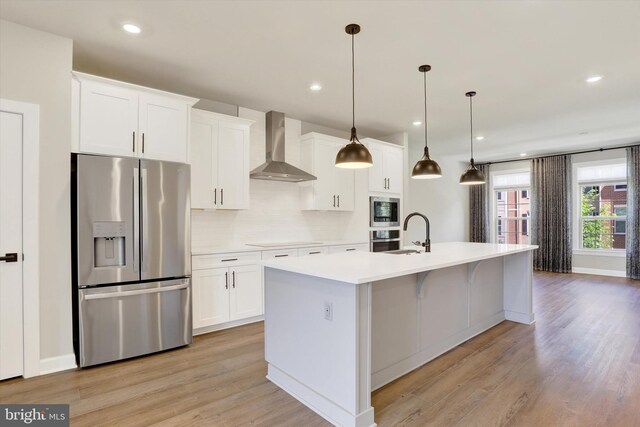 kitchen featuring appliances with stainless steel finishes, wall chimney range hood, a kitchen island with sink, a sink, and decorative backsplash