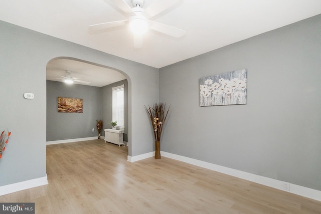 empty room featuring a ceiling fan, arched walkways, light wood-type flooring, baseboards, and radiator heating unit