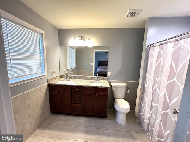 ensuite bathroom featuring tile walls, tile patterned flooring, wainscoting, double vanity, and toilet
