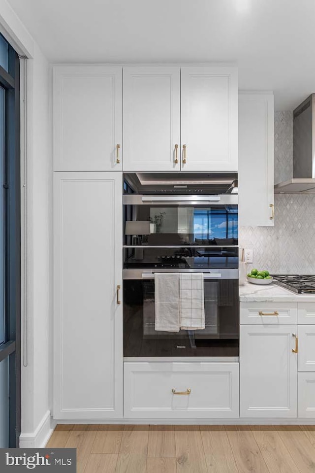 kitchen with wall chimney range hood, stainless steel gas cooktop, double wall oven, and white cabinetry