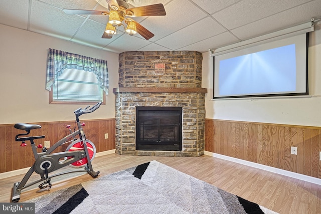 exercise room with a stone fireplace, a drop ceiling, wood finished floors, and a wainscoted wall