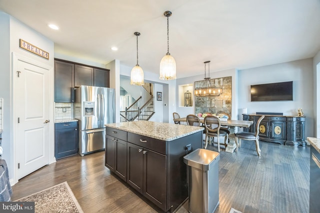 kitchen featuring a center island, stainless steel refrigerator with ice dispenser, light stone countertops, decorative light fixtures, and dark wood-style floors
