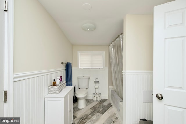 bathroom with toilet, wood finished floors, shower / tub combo with curtain, and a wainscoted wall