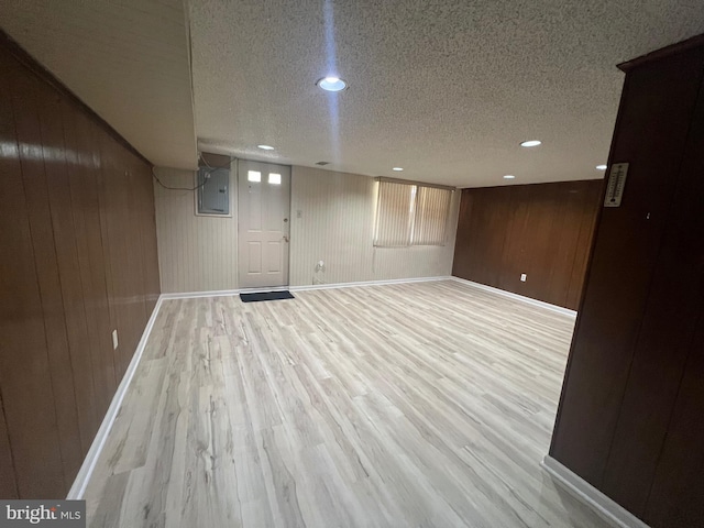 finished basement with wooden walls, a textured ceiling, electric panel, and light wood-type flooring