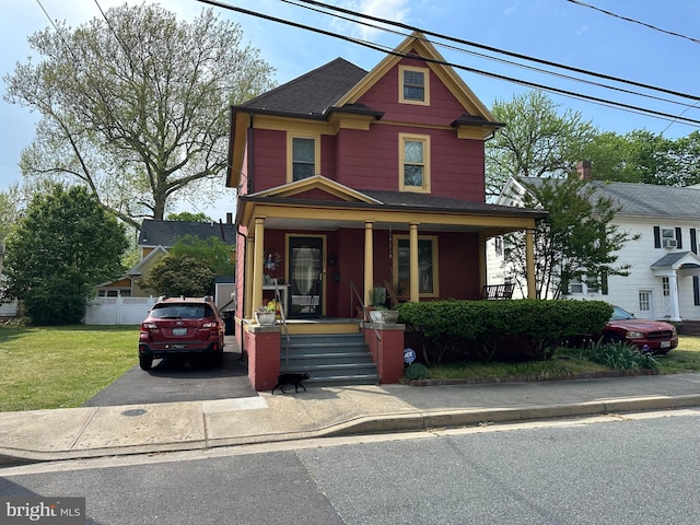 victorian-style house featuring covered porch, fence, a shingled roof, and a front yard