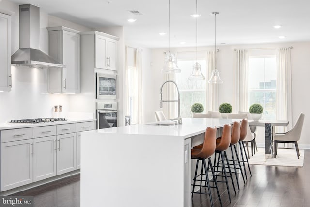 kitchen featuring wall chimney range hood, a sink, a kitchen island with sink, dark wood finished floors, and stainless steel appliances