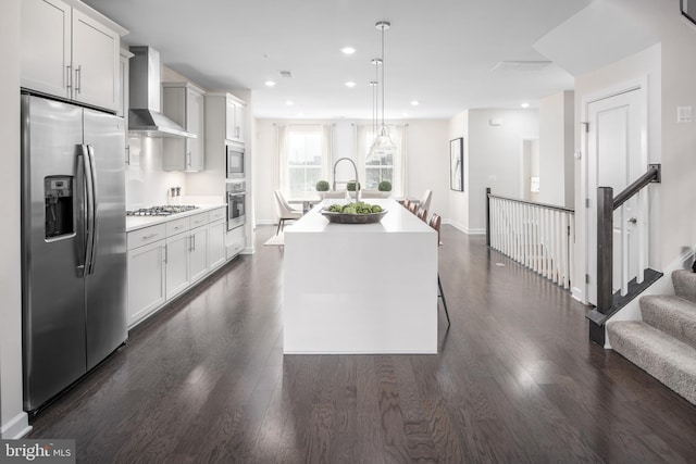 kitchen featuring dark wood-style flooring, a kitchen island with sink, wall chimney range hood, light countertops, and appliances with stainless steel finishes
