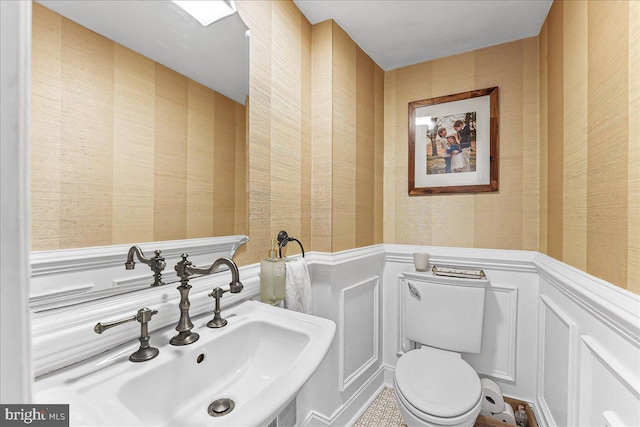 half bath with a sink, wainscoting, toilet, and wallpapered walls