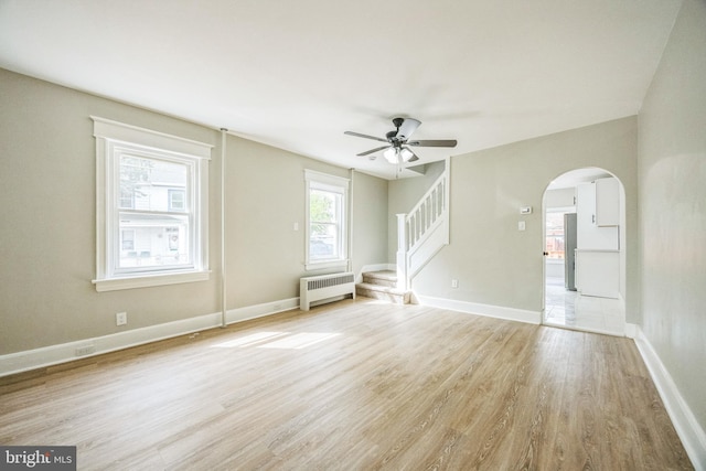 unfurnished living room featuring a ceiling fan, radiator, stairs, arched walkways, and light wood-style flooring