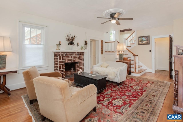 living room featuring ceiling fan, a brick fireplace, light wood-type flooring, baseboards, and stairs