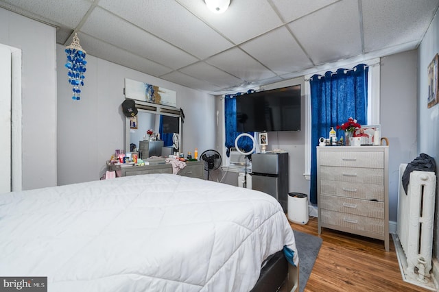 bedroom featuring freestanding refrigerator, radiator heating unit, wood finished floors, and a paneled ceiling