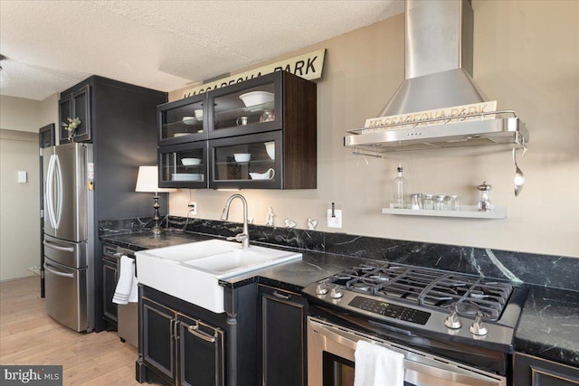 kitchen featuring light wood finished floors, a sink, dark countertops, wall chimney exhaust hood, and appliances with stainless steel finishes