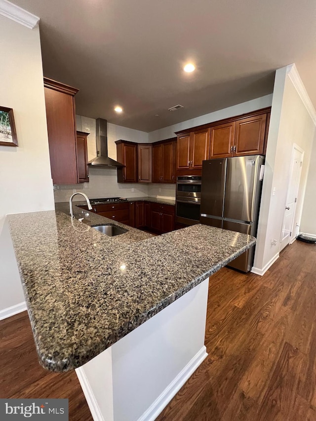 kitchen with wall chimney range hood, appliances with stainless steel finishes, a sink, dark stone countertops, and dark wood-type flooring