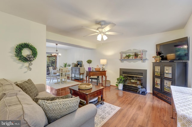 living room featuring baseboards, a fireplace with flush hearth, ceiling fan with notable chandelier, and light wood-type flooring