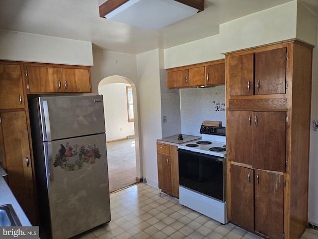 kitchen with brown cabinetry, range with electric cooktop, arched walkways, light countertops, and freestanding refrigerator