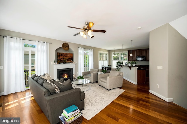 living area featuring ceiling fan, dark wood-style floors, a glass covered fireplace, and baseboards
