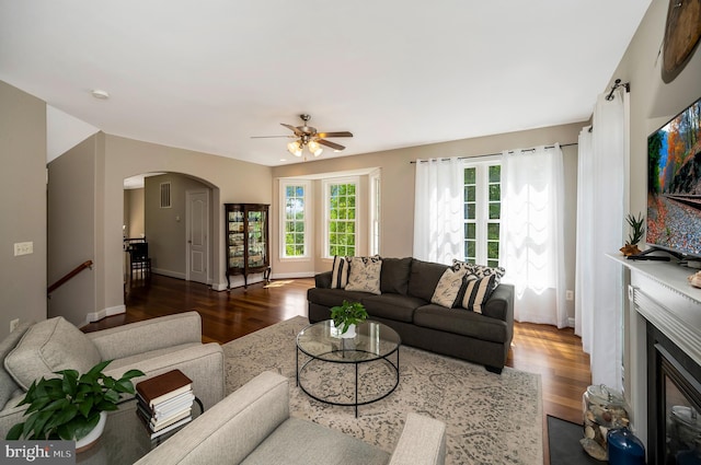 living room featuring wood finished floors, arched walkways, ceiling fan, a glass covered fireplace, and baseboards