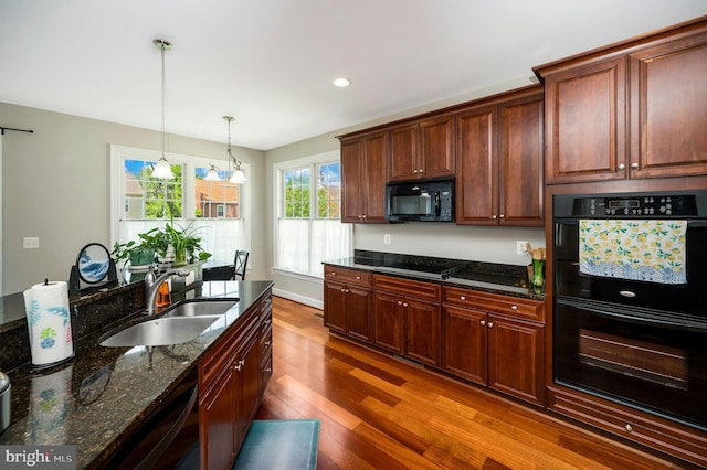 kitchen featuring a sink, black appliances, wood finished floors, pendant lighting, and dark stone counters