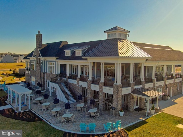 back of property at dusk featuring stone siding, a patio, a chimney, metal roof, and a standing seam roof
