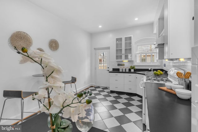 kitchen featuring gas range, white cabinetry, dark countertops, a sink, and glass insert cabinets