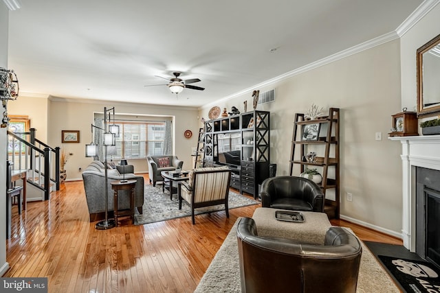 living area with stairs, a ceiling fan, light wood-style flooring, a fireplace with flush hearth, and ornamental molding