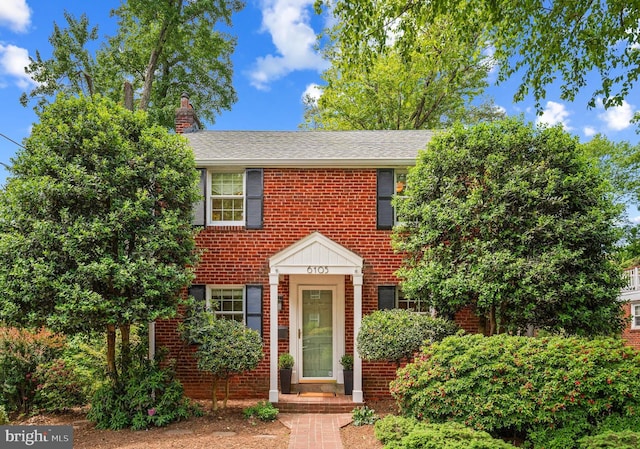 colonial inspired home with brick siding, a chimney, and roof with shingles